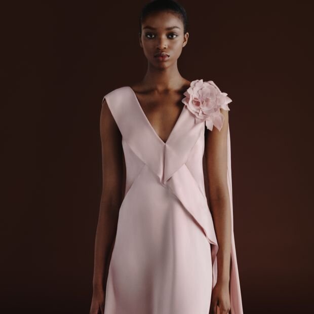 A model wearing a pink satin gown featuring a waterfall drape and a tonal 3D flower appliqué and ruffle details.