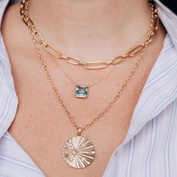 A model wearing 3 gold necklaces layered, a chain necklace, an aquamarine pendant, and a medallion necklace with sapphire stone details.