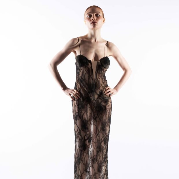 A model wearing a neutral toned bustier maxi dress with sheer black Chantilly lace appliqué and a sculpted bodice.