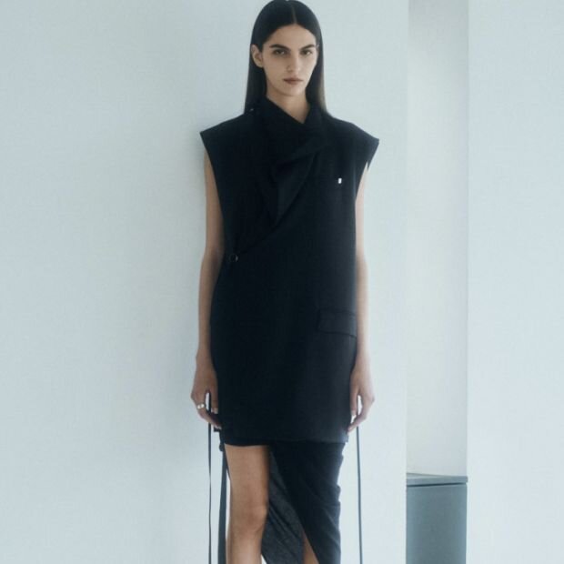 A model wearing a black mini tailored wrap dress, featuring a high collar that unbuttons to reveal a draped cowl neckline