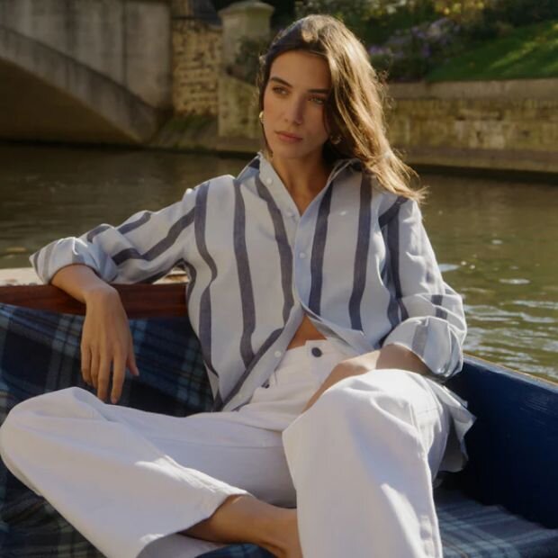 A model sitting on a rowboat in a white button-up shirt with grey stripes, paired with relax-fitted white jeans.