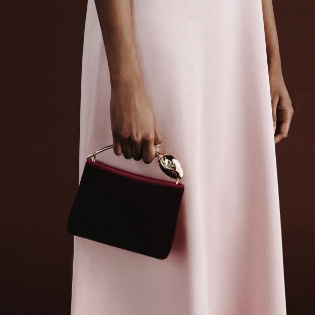 A model carrying a mini red velvet rectangular bag with a goldtone flower bud handle and a petal-shaped magnetic closure.