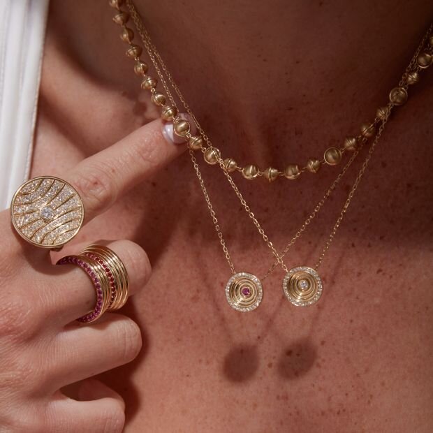 A model wearing a gold necklace layered with 2 pendant necklaces, with multiple cabochon and band rings.