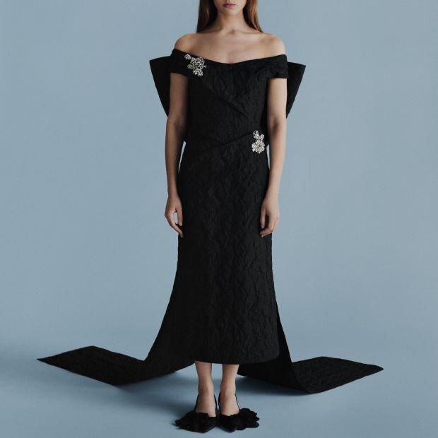 A model wearing a black cloqué jacquard off-shoulder midi dress featuring an oversized, trailing tulle bow, with crystal embroideries at the neckline and hip.