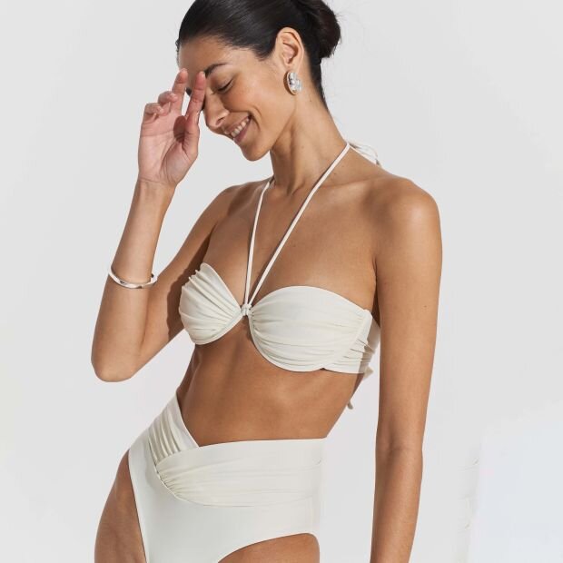 A model wearing a white bikini top with a balconette bust with ruched cups and a back-tie closure, with a matching high-waisted bikini bottom.