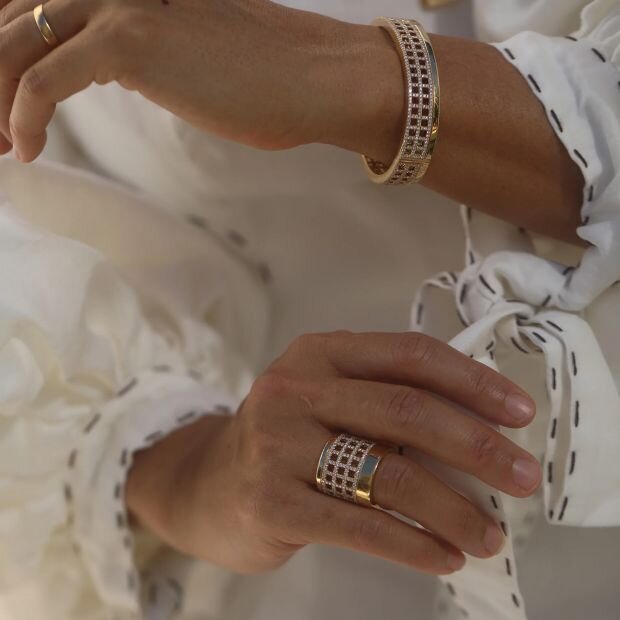 A model wearing a 18k gold diamond ring featuring a wide band with an open-worked, grid-like pattern. Paired with a matching bracelet.