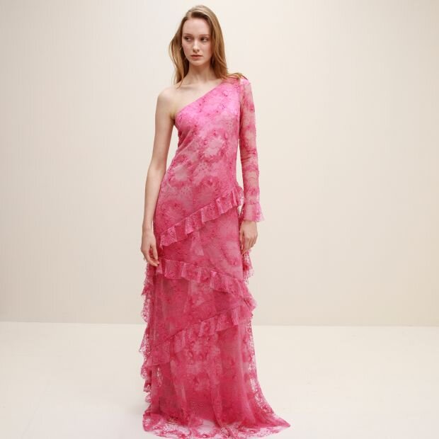 A model wearing a pink asymmetric maxi dress with an embroidered lace silhouette and ruffle embellishments, featuring a slanted neckline and single long sleeve.
