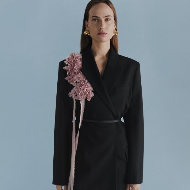 A model wearing a black wool double-breasted blazer with a waist-cinching tie and blush pink silk ribbon appliqué flowers.
