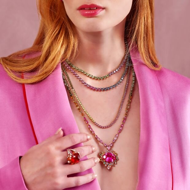 A woman wearing a red ruby ring and a gold band with gemstone details, and a pendant necklace featuring multiple rainbow-colored gemstones