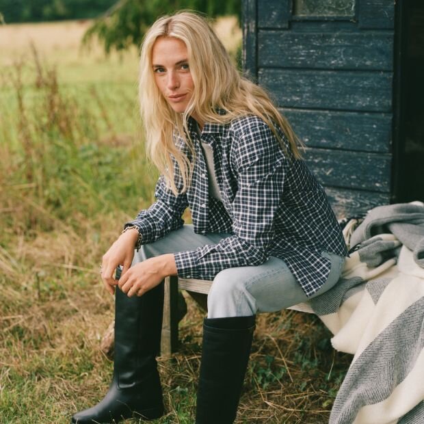 A model wearing a black and white plaid button up over a knit, paired with faded blue jeans and riding boots.