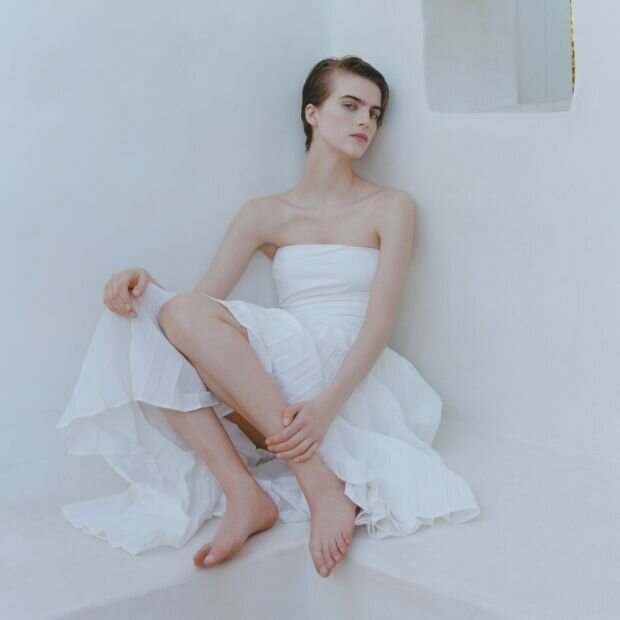A woman wearing a white strapless midi dress featuring a ruched bustier bodice with a pleated poplin skirt.