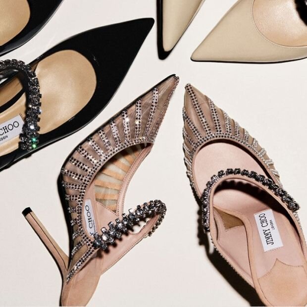 A pair of crystal-embellished suede mesh mule in nude with a stiletto heel and pointed toe, and the mules in a black colorway next to them.