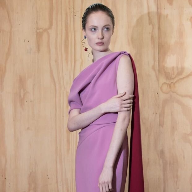 A model wearing a pink crepe midi dress with an  asymmetrically folded neckline with a trailing cape at the opposite shoulder.