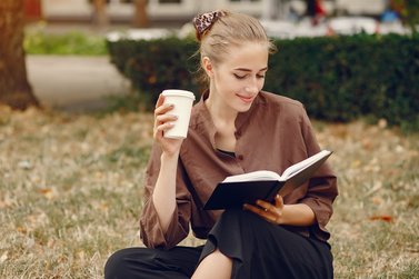 woman sitting on the grass smiling while reading a book and drinking a hot beverage
