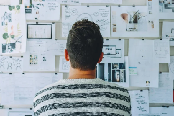 Man in a striped sweater reviewing a wall filled with notes, sketches, and brainstorming diagrams, symbolizing planning or strategy development.