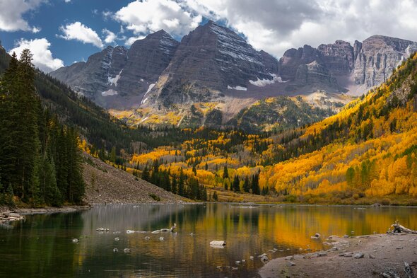 cenic view of snow-capped mountains, surrounded by autumn trees reflecting on a tranquil lake, symbolizing the beauty of Colorado's natural landscape