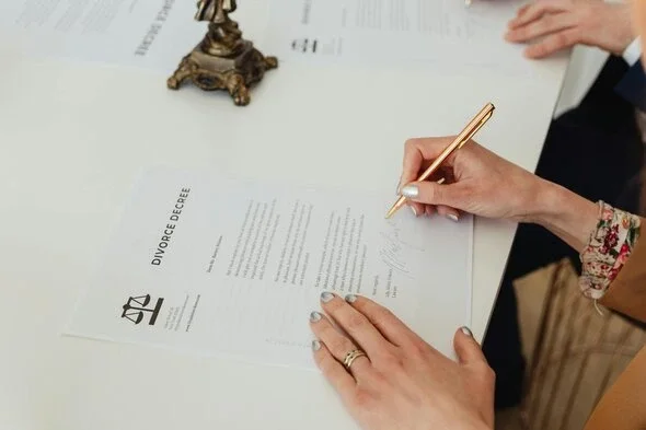 A close-up of a person's hands signing a divorce decree document with a gold pen on a white table.