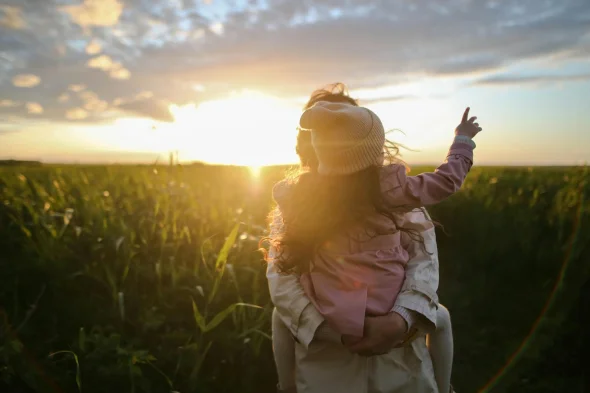 A parent holding a child in a field, pointing at the sunset.