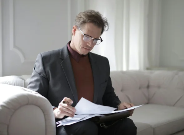 A man in a suit sitting on a sofa, reviewing documents attentively.