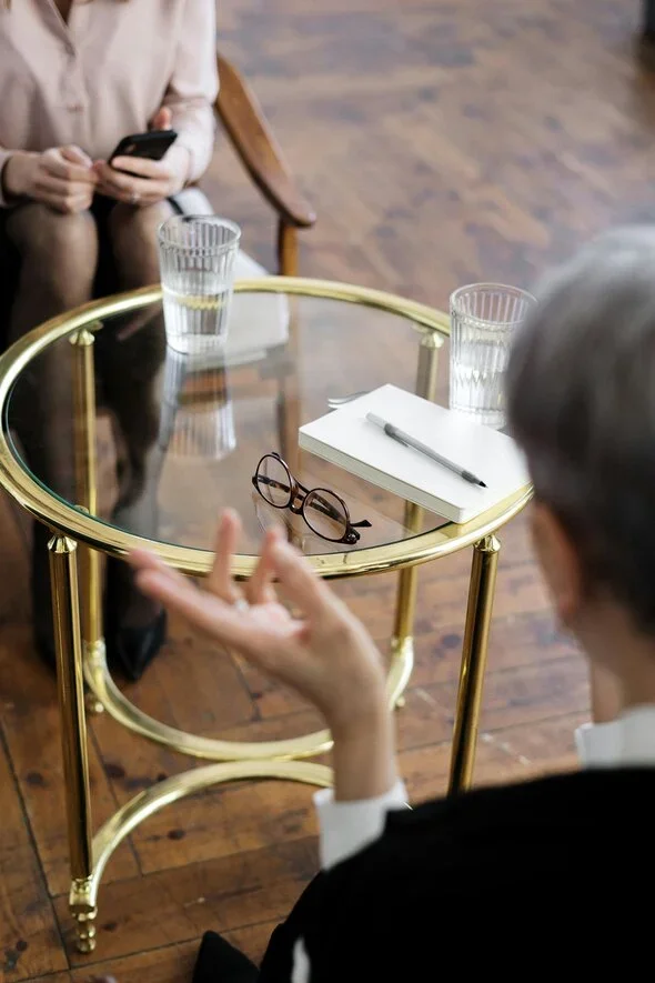 Two individuals in a formal setting sitting across from each other with glasses of water and notes on a round glass table, symbolizing a legal discussion or counseling session.