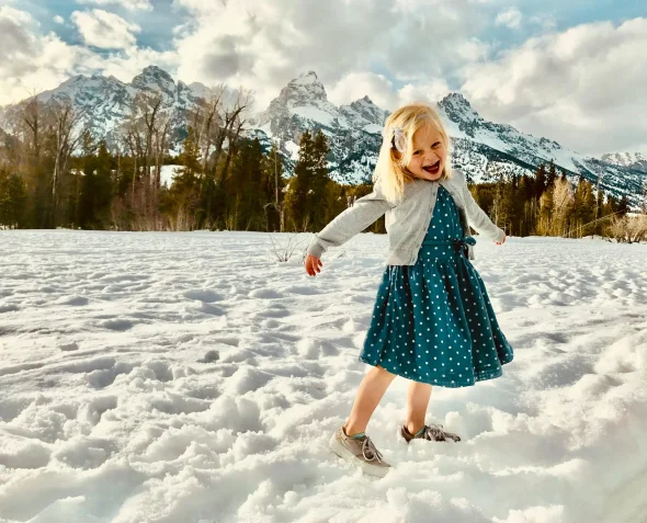 A joyful young girl playing in the snow, with snowy mountains and trees in the background, capturing the carefree spirit of childhood in a scenic, wintry landscape.