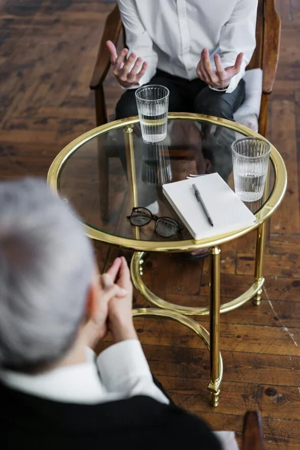 Two individuals in a formal discussion setting, seated across from each other with glasses of water and notes on a glass table, symbolizing legal consultation or negotiation.