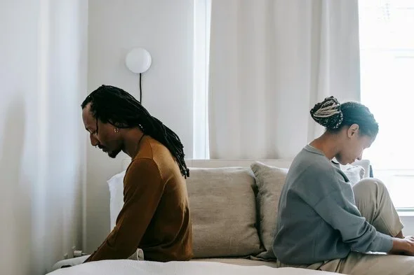 A couple sitting back-to-back on a bed, each looking down with a sense of distance, illustrating emotional tension and contemplation.