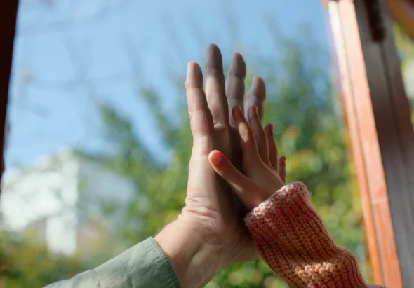 A child and his parent holding hands through a glass window