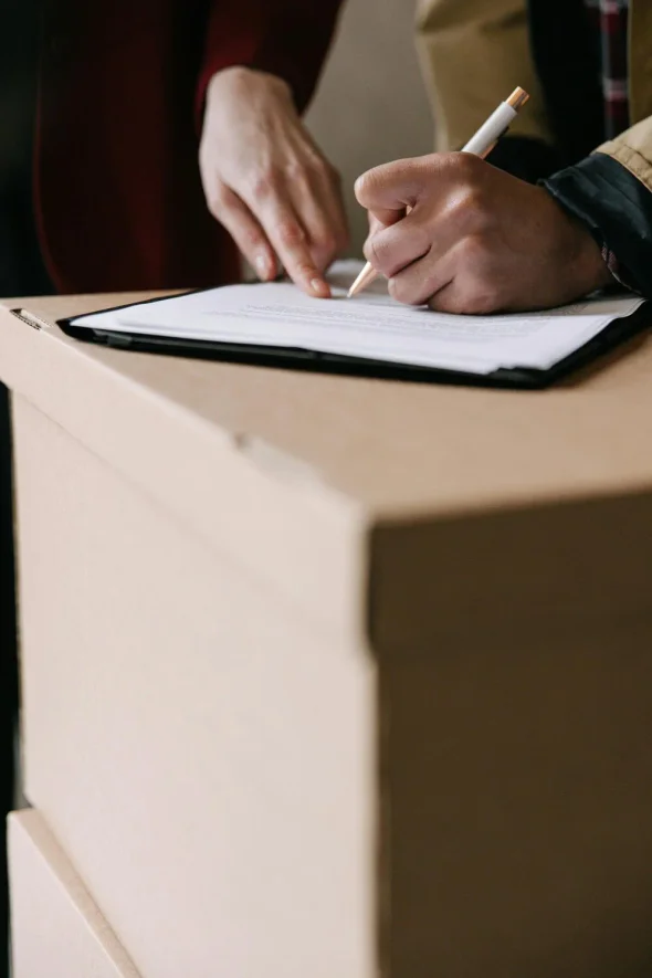 A person signing a document on a clipboard placed on cardboard boxes.