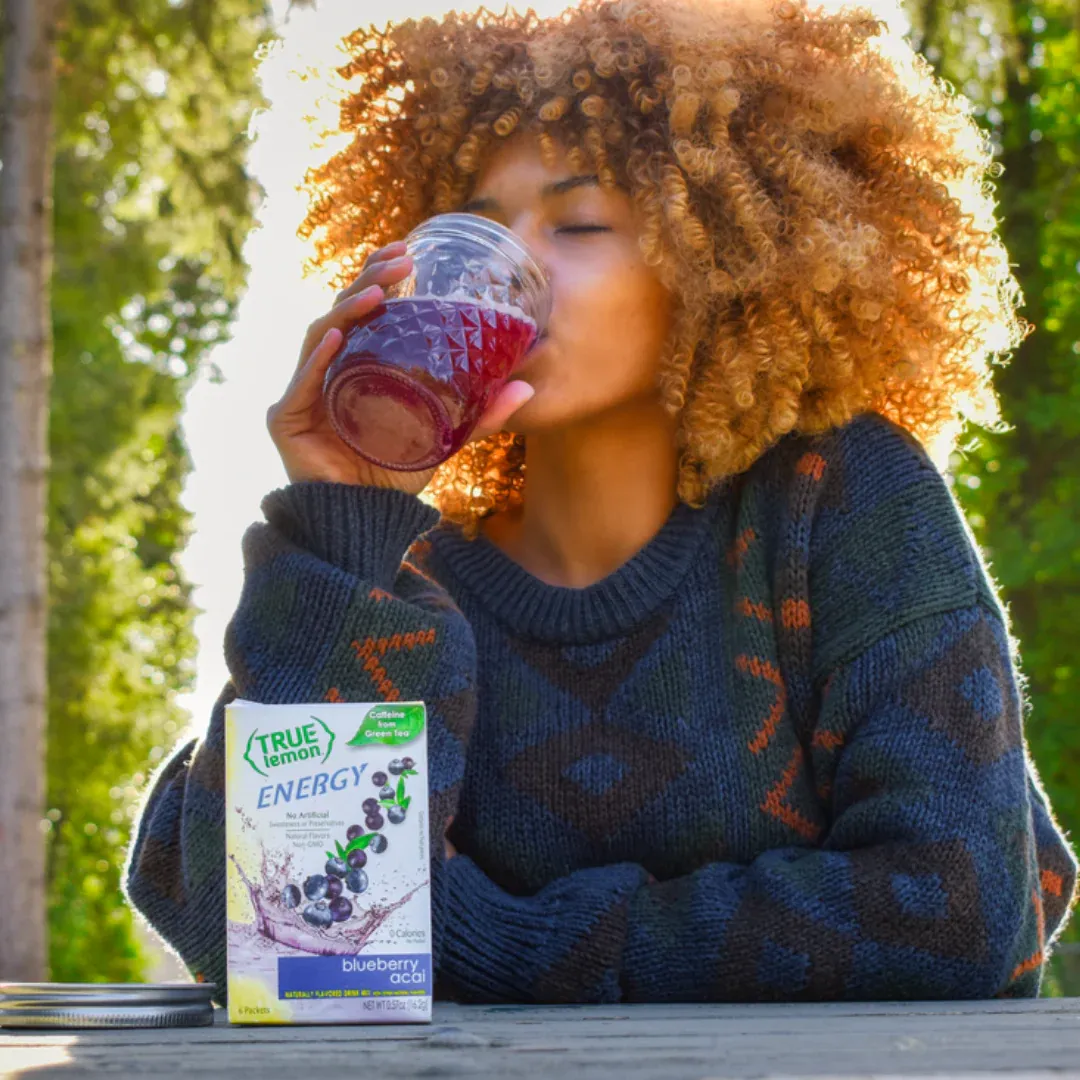 A woman sitting outdoors drinking a glass of True Lemon Energy Blueberry Acai.