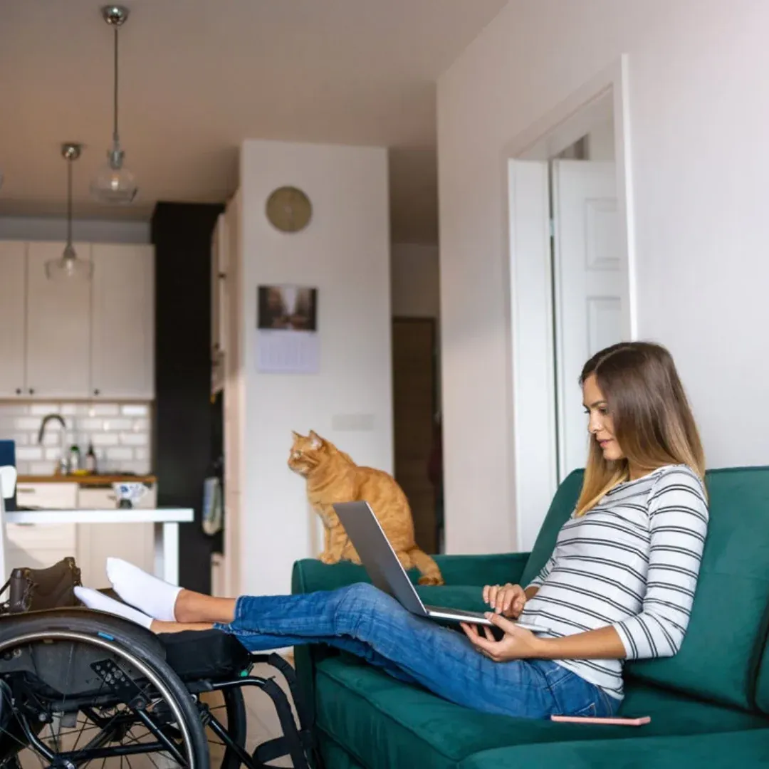 A woman is sitting on her couch next to a cat, she is looking at her laptop.