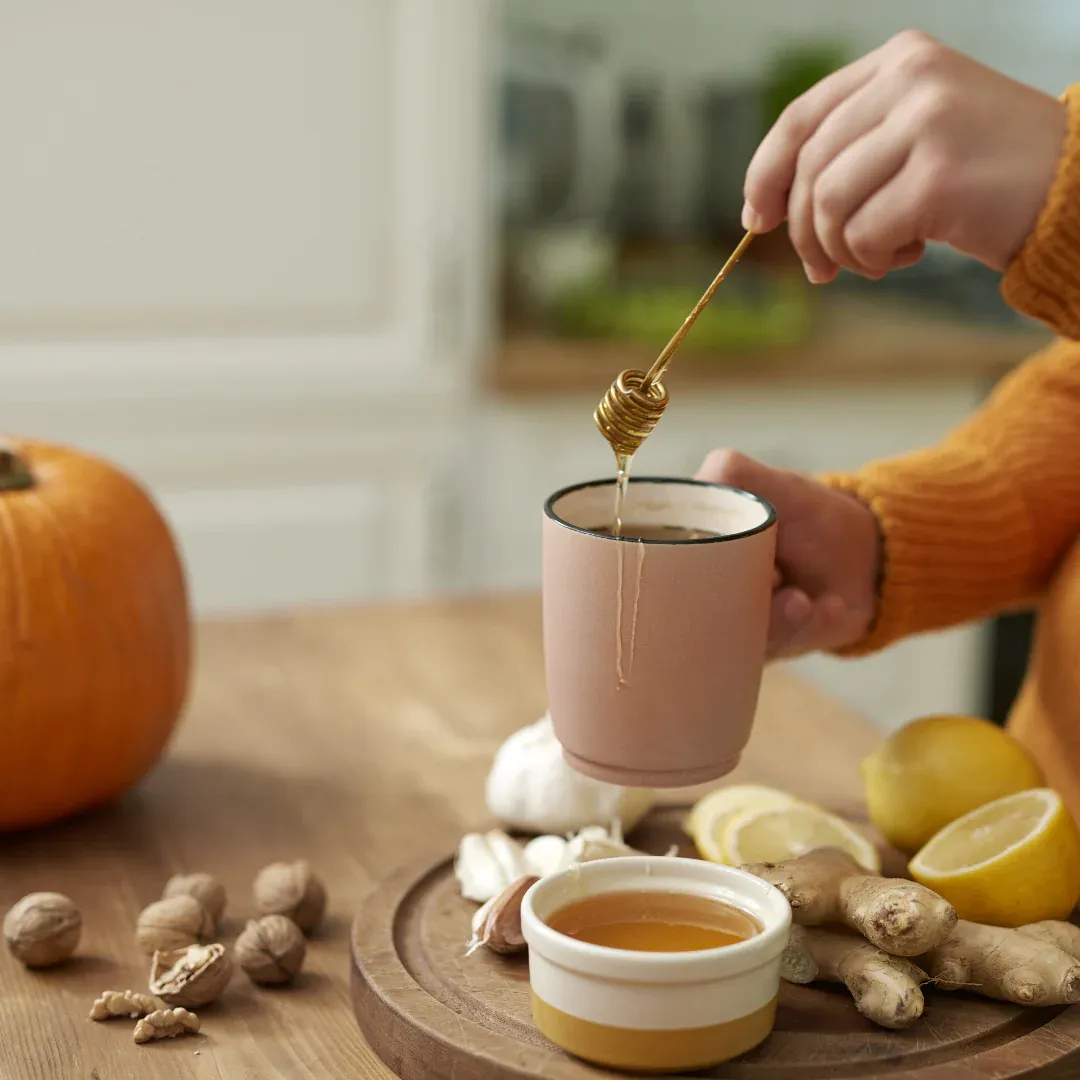 A person in the kitchen putting honey in a mug of tea. There are other ingredients on the counter.