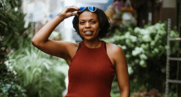 A woman in red tank top is smiling. She is standing in front of plants.