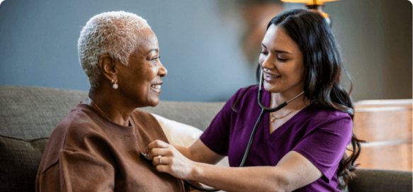 Healthcare worker using a stethoscope to examine a smiling senior woman's heart in a home setting