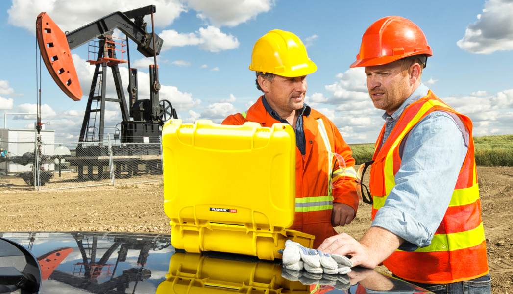 2 workers wearing PPE are looking at something in a yellow Nanuk case. An oil well is in the background
