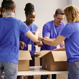 Four volunteers wearing matching purple shirts and lanyards pack canned goods into cardboard boxes.