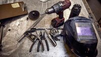 Assorted hand tools and equipment, including a cordless drill, pliers, measuring tools, and a welding helmet, arranged on a metal workbench.