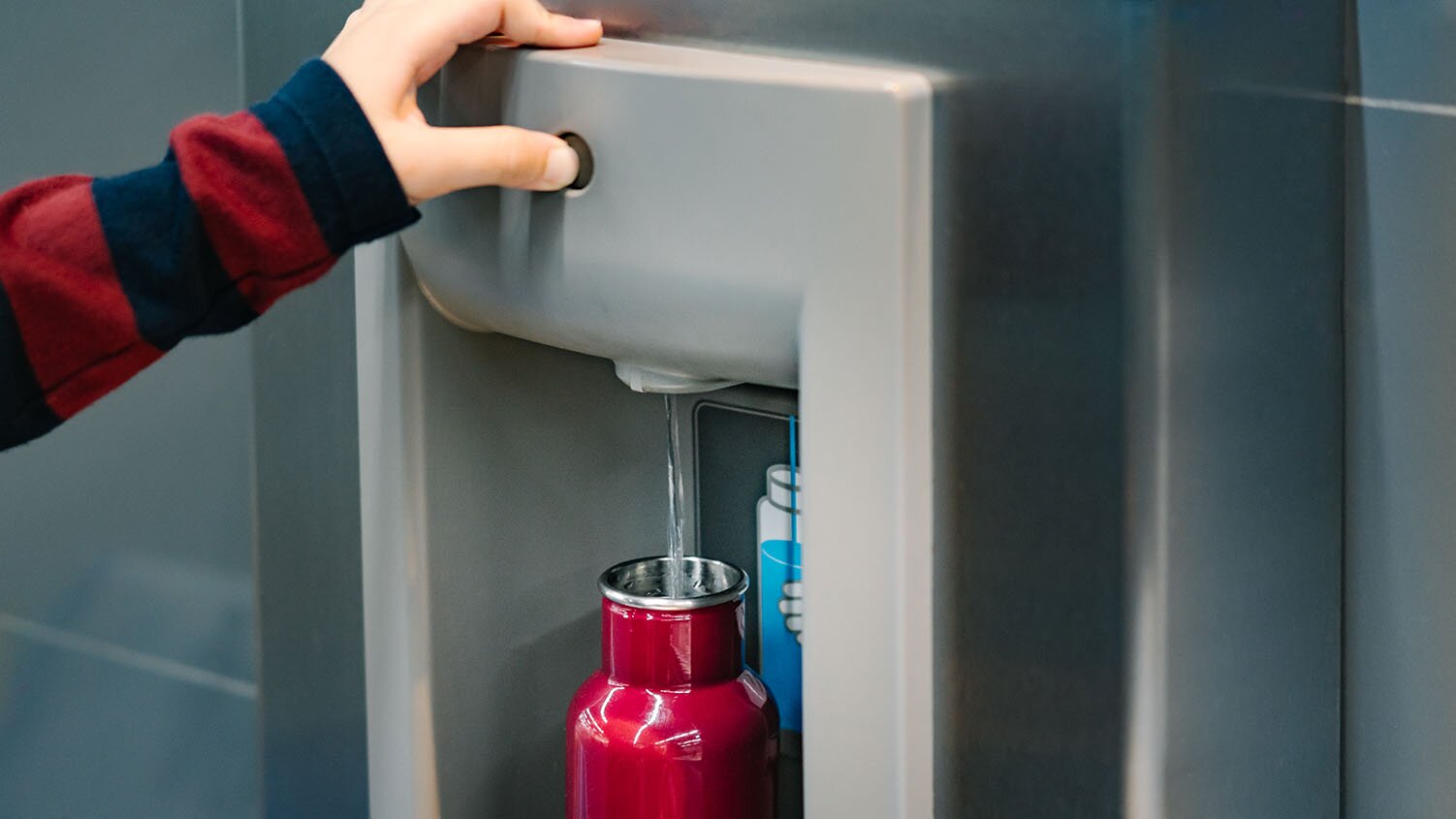 Image of a thumb pressing a button on a water filling station. Water is flowing into a water bottle.