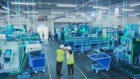 Two workers in safety vests standing inside a modern manufacturing facility with digital interface graphics overlaying automated production equipment.