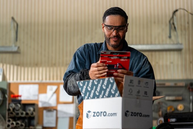 Man wearing safety glasses and dark blue work shirt holding red handheld device or scanner while standing in industrial warehouse or manufacturing facility with corrugated metal walls and machinery in background