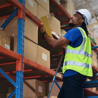 Worker in a hard hat and high-visibility vest stands on a step ladder while inspecting boxed inventory in a warehouse.