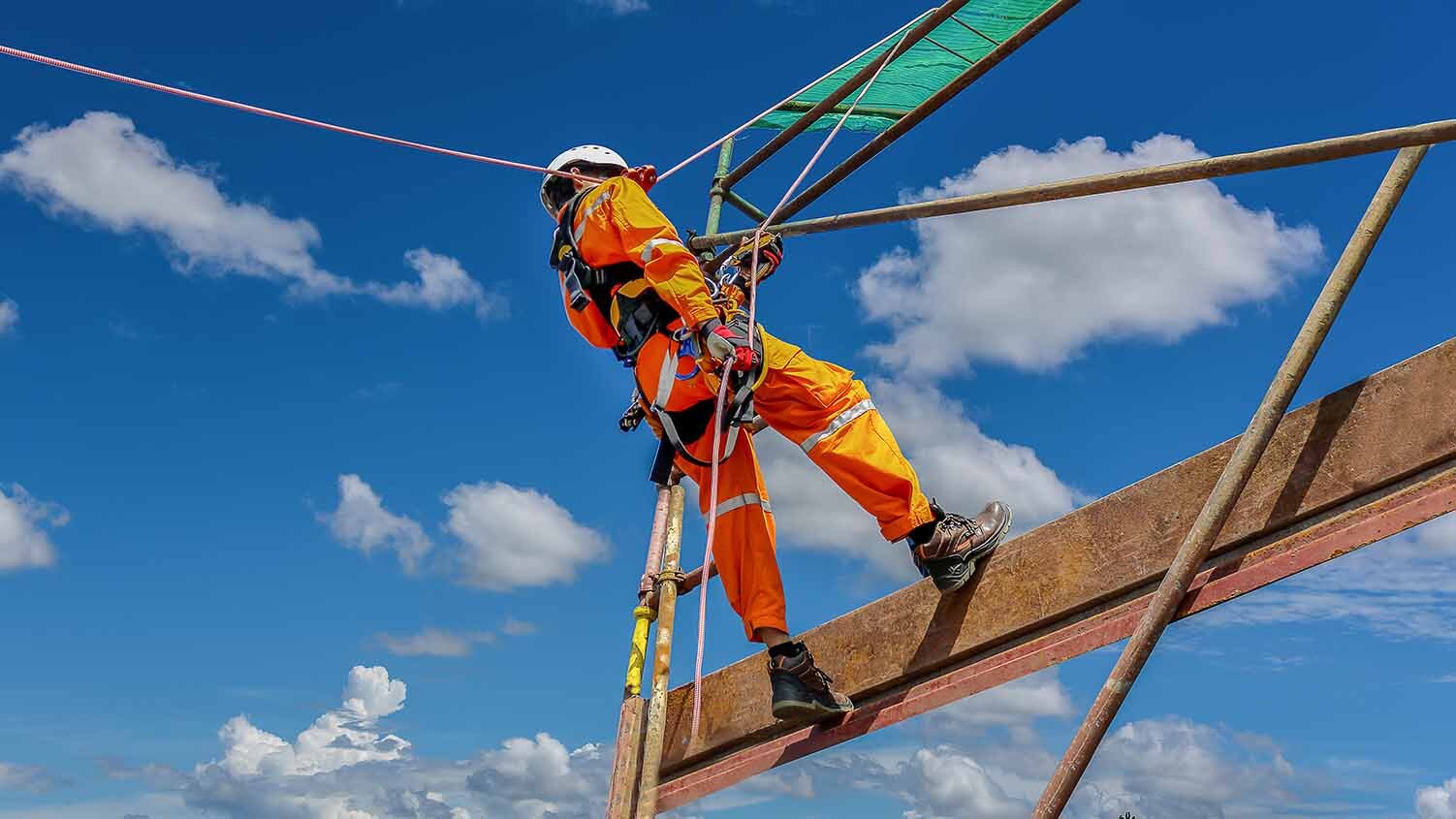 Image of a worker in an orange jumpsuit and hard hat wearing a full-body harness and dangling off scaffolding.
