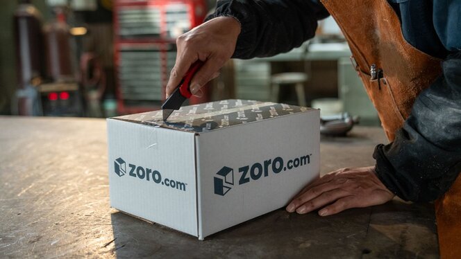 Warehouse worker using a box cutter to open a Zoro-branded shipping box on a workbench.