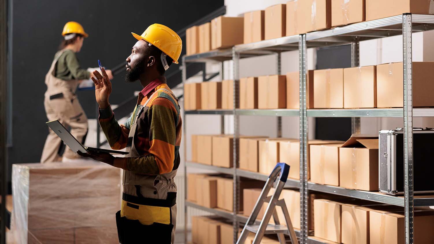 Image of a man wearing protective overalls and a hard hat. He is standing in a warehouse holding a laptop and ballpoint pen, and appears to be counting inventory. Behind him are shelves full of brown boxes, and other boxes are shrink-wrapped together on a pallet.