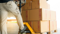 Worker using a pallet jack to move stacked cardboard boxes labeled with handling symbols in a warehouse setting.
