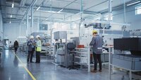Modern manufacturing floor with workers in safety gear operating machinery and reviewing a clipboard in a clean, well-lit industrial facility.