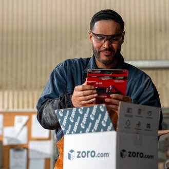 Warehouse employee wearing safety goggles checks a packaged item beside an open Zoro.com shipping box.