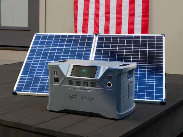 A Patriot Power Generator 2000x sitting outside on someone's porch charging with its solar panel, and an American Flag hanging on the wall