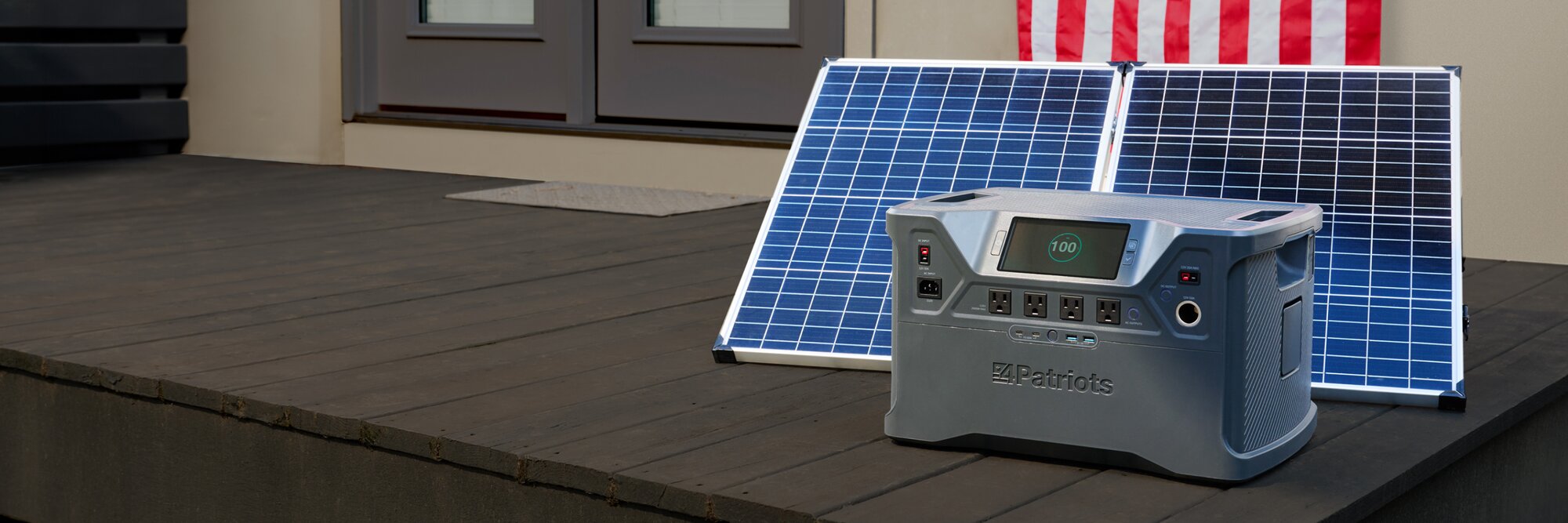 A Patriot Power Generator 2000x sitting outside on someone's porch charging with its solar panel, and an American Flag hanging on the wall