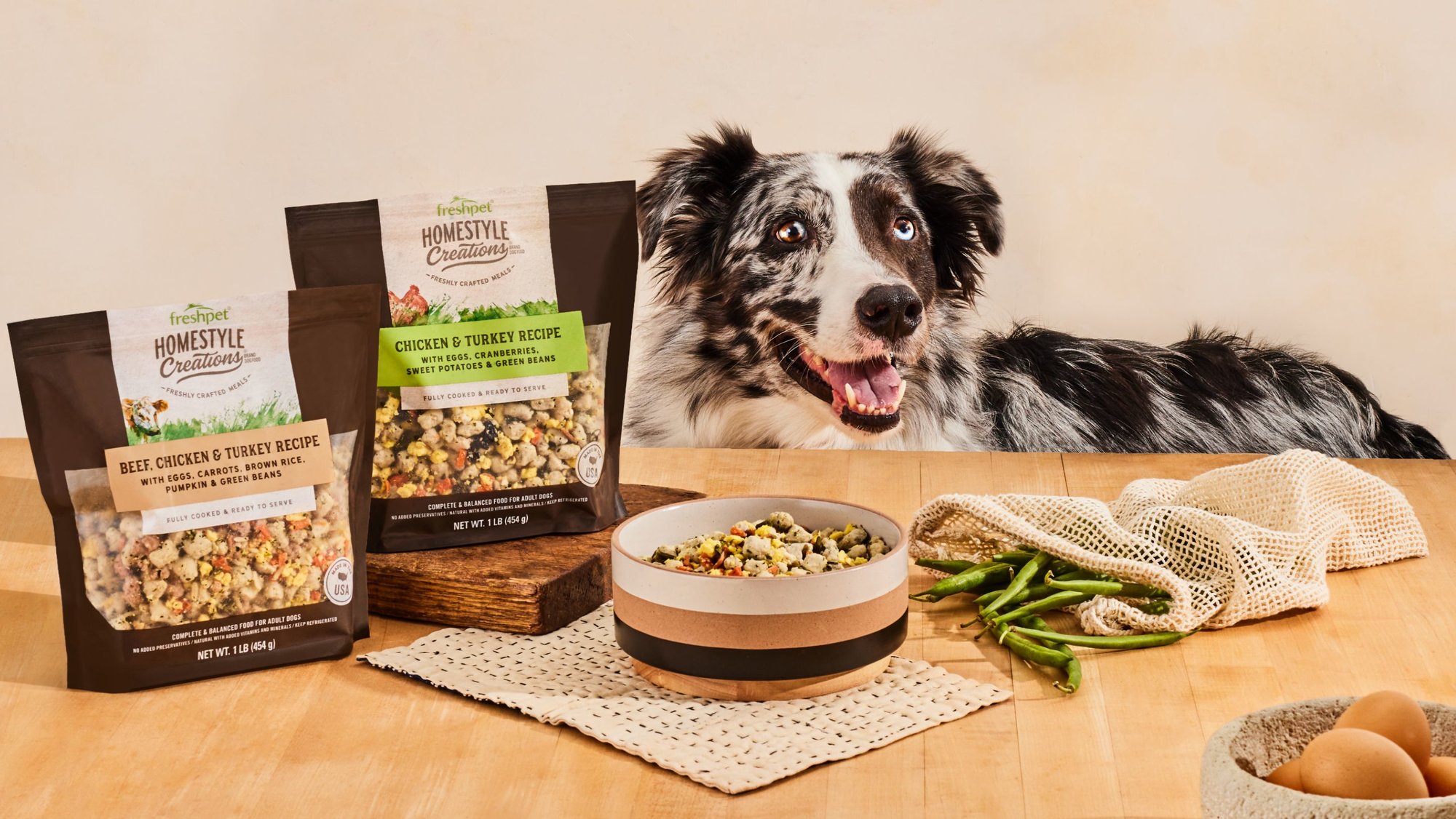 A black and white dog behind a bag and bowl containing Freshpet Homestyle Creations recipe.